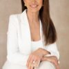 Mature woman with long brown hair smiling confidently while wearing a smart white blazer and gold jewellery.