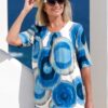vanessa-crookes-20 A mature woman in a patterned blue top and hat smiles on a sunny seaside holiday balcony.
