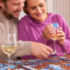 Couple Sitting At Table At Home With Glass Of Wine Doing Jigsaw Puzzle Together
