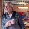 Portrait Of Senior Male Wearing Overalls In Garage Workshop With Hot Drink