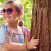 Smiling Young Girl Wearing Sunglasses Playing Hide And Seek Behind Tree In Garden