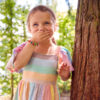 Smiling Young Girl Playing Hide And Seek Behind Tree In Garden