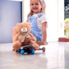 Smiling Young Girl Playing Game Sitting On Skateboard With Teddy Bear At Home
