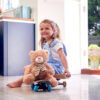Smiling Young Girl Playing Game Sitting On Skateboard With Teddy Bear At Home