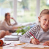 Portrait Of Girl Sitting At Table Home Schooling During Health Pandemic With Mother In Background