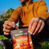 A smiling man sits on the grass outdoors, enjoying a snack of Western Cape beef biltong outdoors.