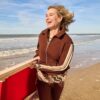 A mature woman smiling joyfully while walking along a sunny beach carrying a red wooden chair.