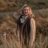 A smiling blonde woman wearing a warm gilet stands in coastal grassy dunes during the golden hour.