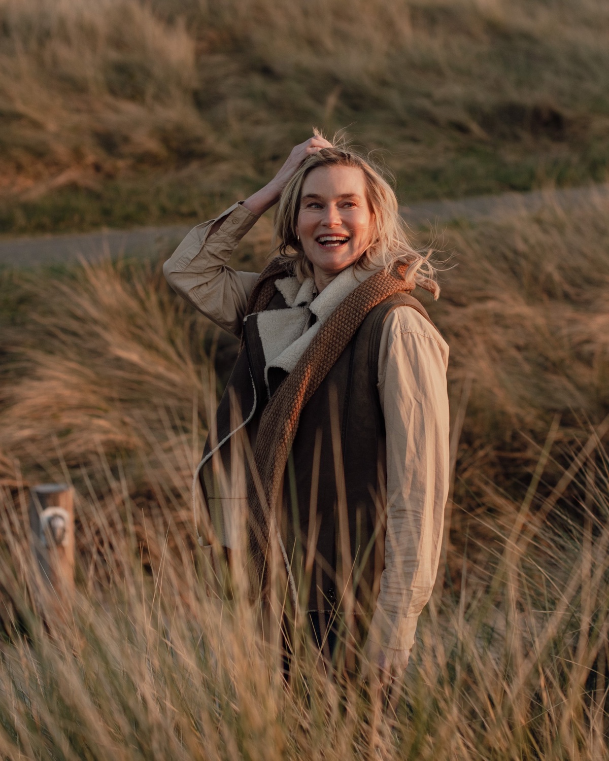 A smiling blonde woman wearing a warm gilet stands in coastal grassy dunes during the golden hour.