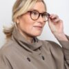 A mature blonde woman wearing stylish tortoiseshell glasses and a beige trench coat looks away thoughtfully.