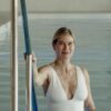A mature blonde woman in a white swimsuit stands in a swimming pool holding a metal rail.