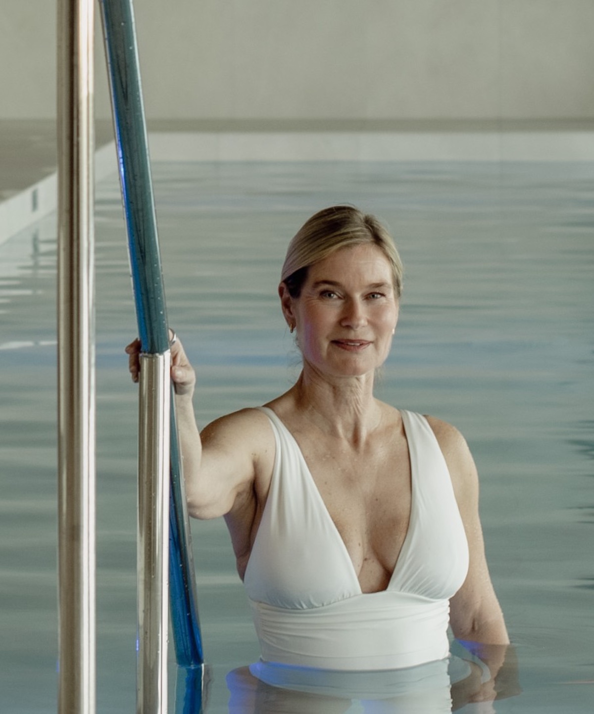 A mature blonde woman in a white swimsuit stands in a swimming pool holding a metal rail.