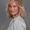 Mature blonde woman with a friendly, professional smile poses against a neutral grey studio backdrop.