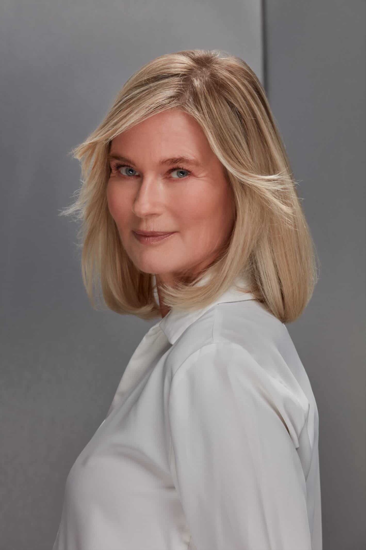 Mature blonde woman with a friendly, professional smile poses against a neutral grey studio backdrop.