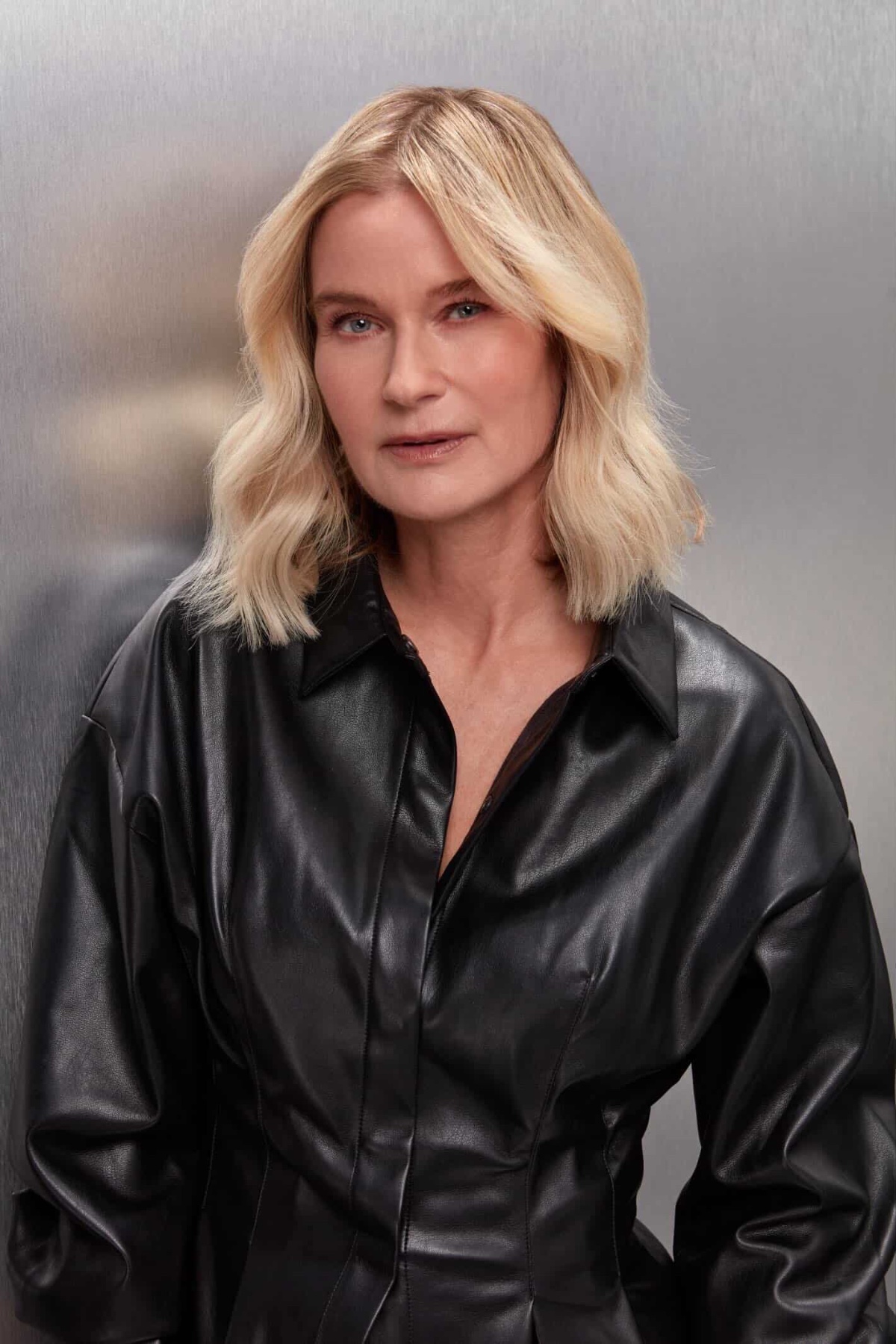 Mature blonde female model with a neutral expression wearing a black leather shirt against metallic background.