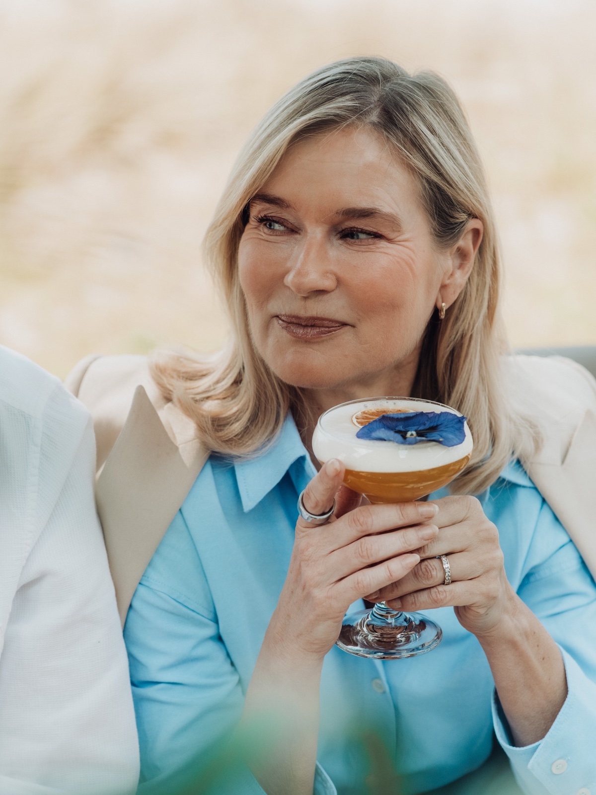 A mature blonde woman holds an elegant cocktail glass while smiling during a relaxed outdoor social afternoon.
