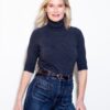A smiling blonde woman wearing a charcoal turtleneck and denim jeans stands against a clean white backdrop.