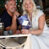 A mature couple sits together at an outdoor cafe table, smiling while looking at a drinks menu.