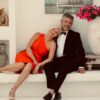A stylish mature couple posing together on a white bench against a bright, elegant Mediterranean-style background.