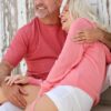A happy middle-aged couple in casual clothing laughing together outdoors against a white wooden wall.