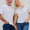 A smiling mature couple in white t-shirts and jeans hold hands against a plain light background.