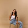 trinh-nguyen-8 A smiling young woman with long dark hair sits on a metal chair against a beige studio backdrop.