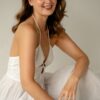 A smiling brunette woman with freckles wearing a white summer dress poses against a neutral studio background.