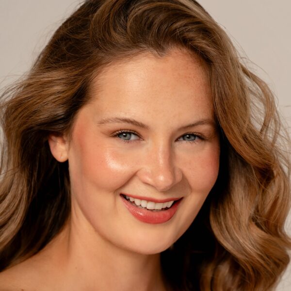 A friendly young woman with wavy brown hair smiles warmly at the camera against a neutral background.