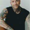 A tattooed, bald man with a friendly smile poses against a neutral background for a commercial portrait.