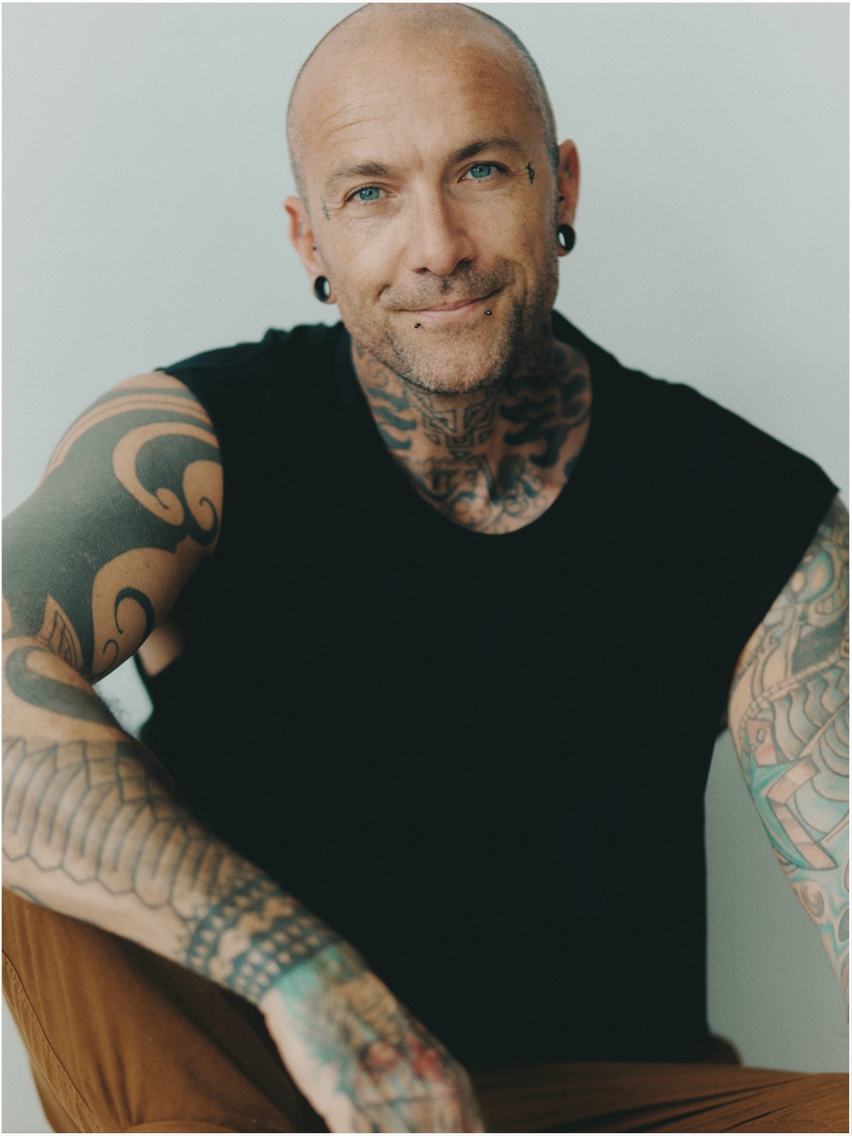 A tattooed, bald man with a friendly smile poses against a neutral background for a commercial portrait.