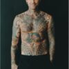 A tattooed bald man smiles warmly while standing against a solid black background in a studio portrait.