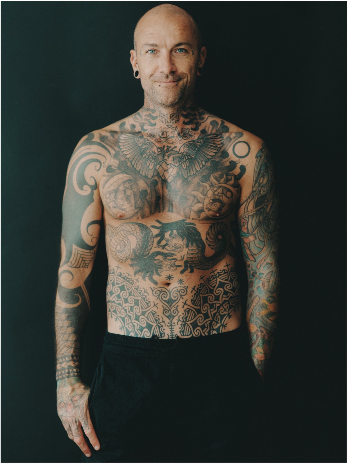 A tattooed bald man smiles warmly while standing against a solid black background in a studio portrait.