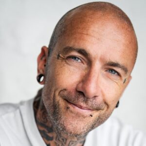 A kind-looking, tattooed man with a bald head and facial piercings smiling warmly at the camera lens.