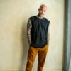 A tattooed man stands barefoot against a textured wall wearing a sleeveless black shirt and orange chinos.