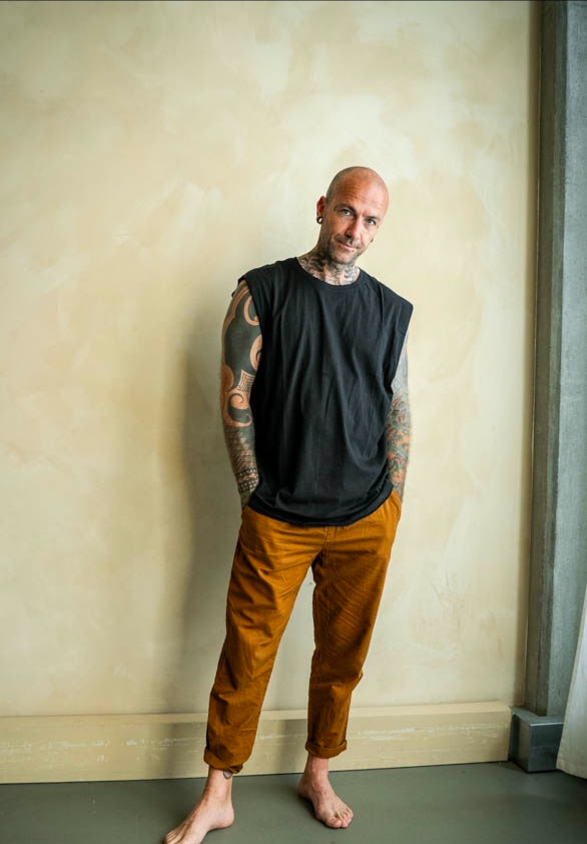 A tattooed man stands barefoot against a textured wall wearing a sleeveless black shirt and orange chinos.