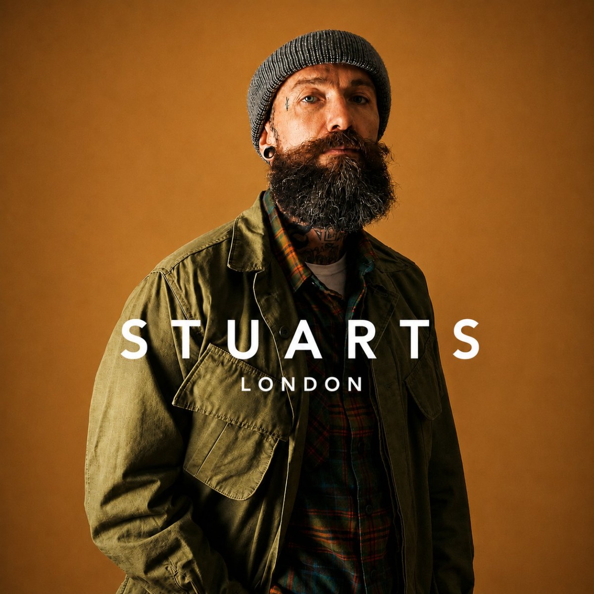 A bearded man with tattoos wearing a beanie and olive jacket poses against a mustard yellow studio background.
