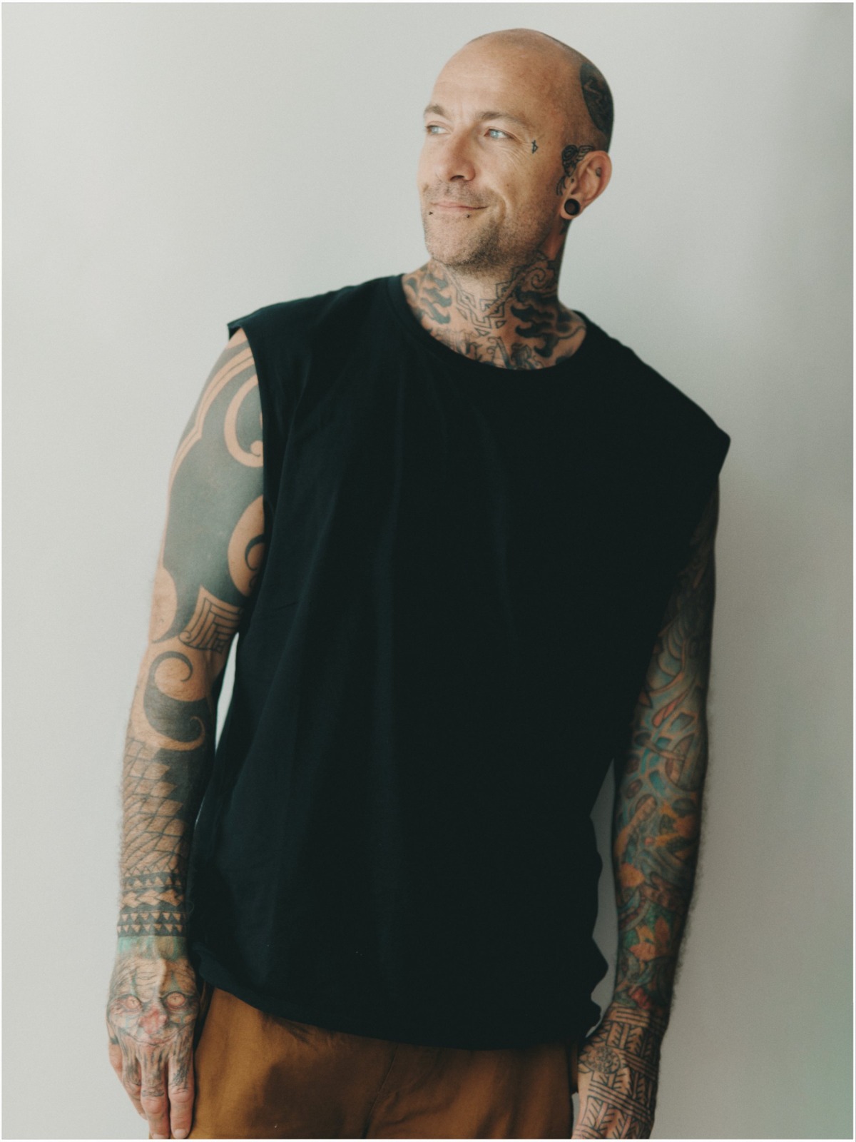 A bald, heavily tattooed man with a friendly smile looks away from the camera against a plain background.