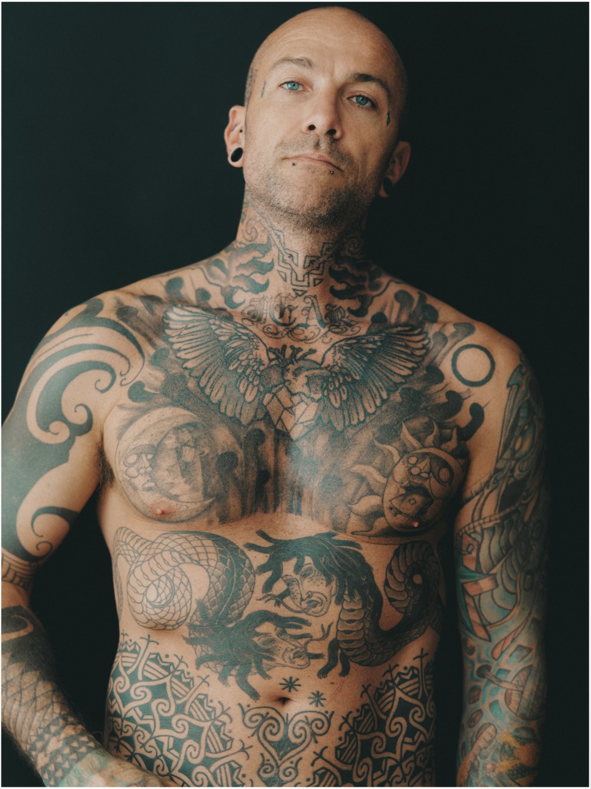 A bald, heavily tattooed man with body art pose against a simple black background for a portrait.