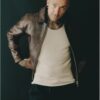 A tattooed bald man in a leather jacket and beige vest poses against a dark studio background.