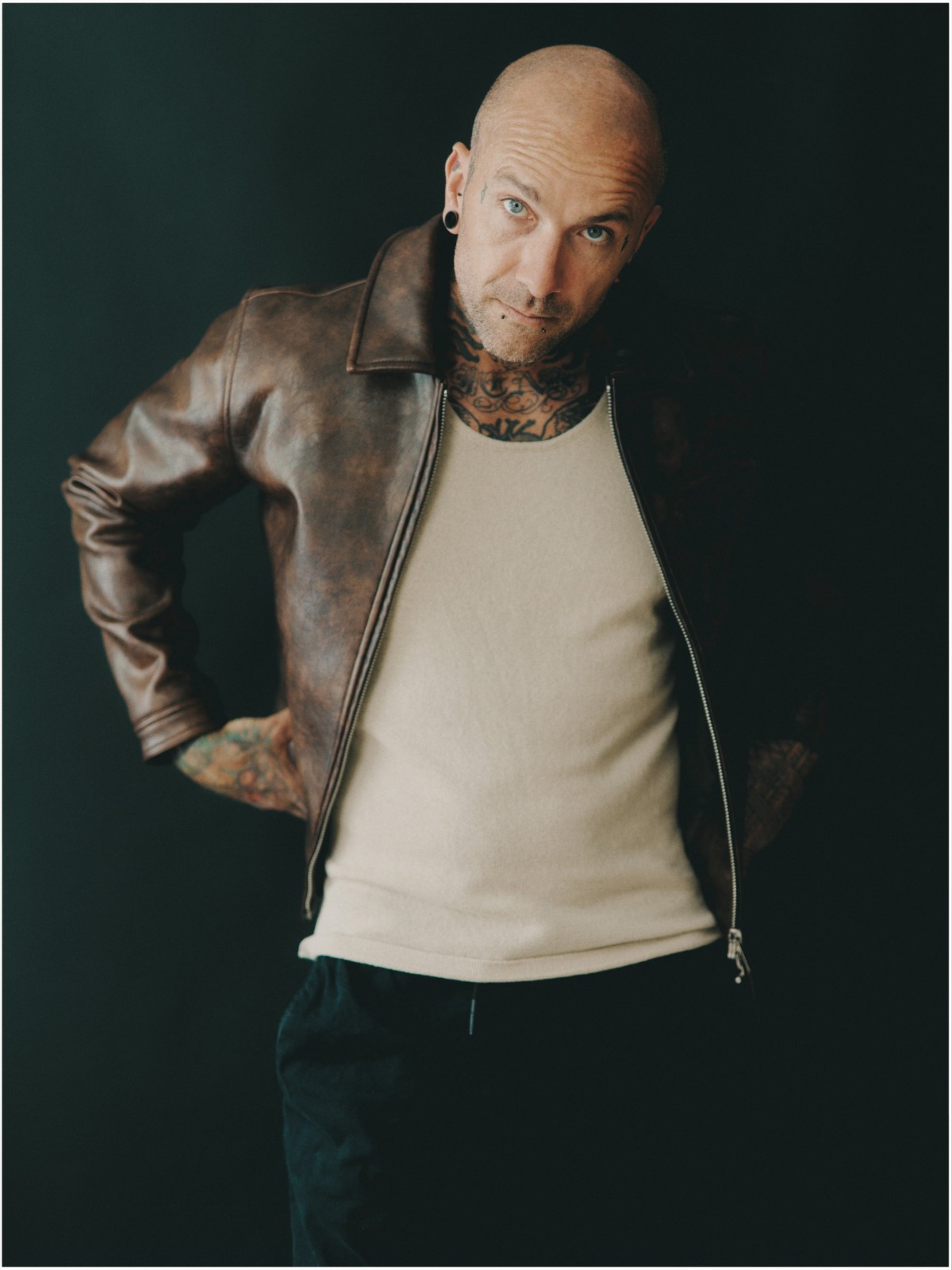 A tattooed bald man in a leather jacket and beige vest poses against a dark studio background.