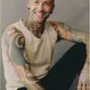A tattooed man with a friendly smile wearing a beige vest against a soft grey studio background.
