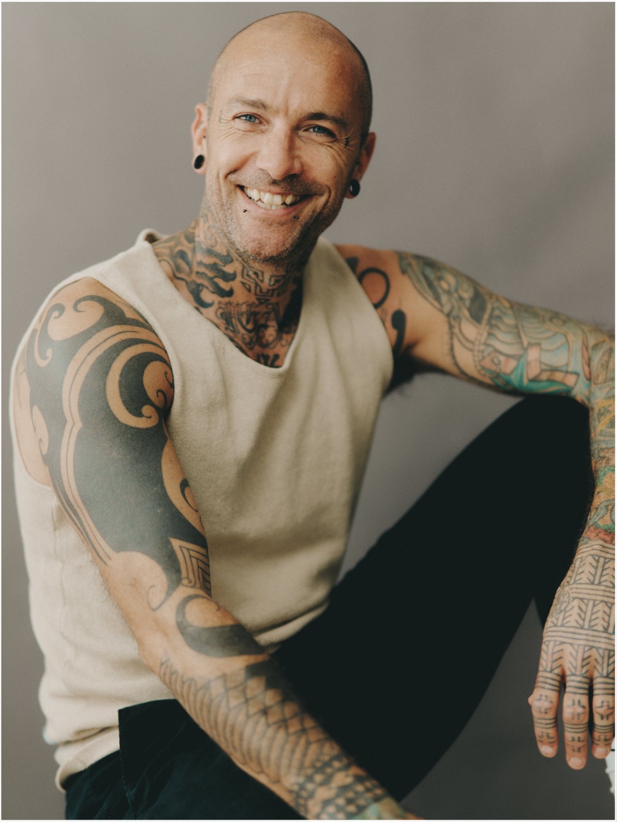 A tattooed man with a friendly smile wearing a beige vest against a soft grey studio background.