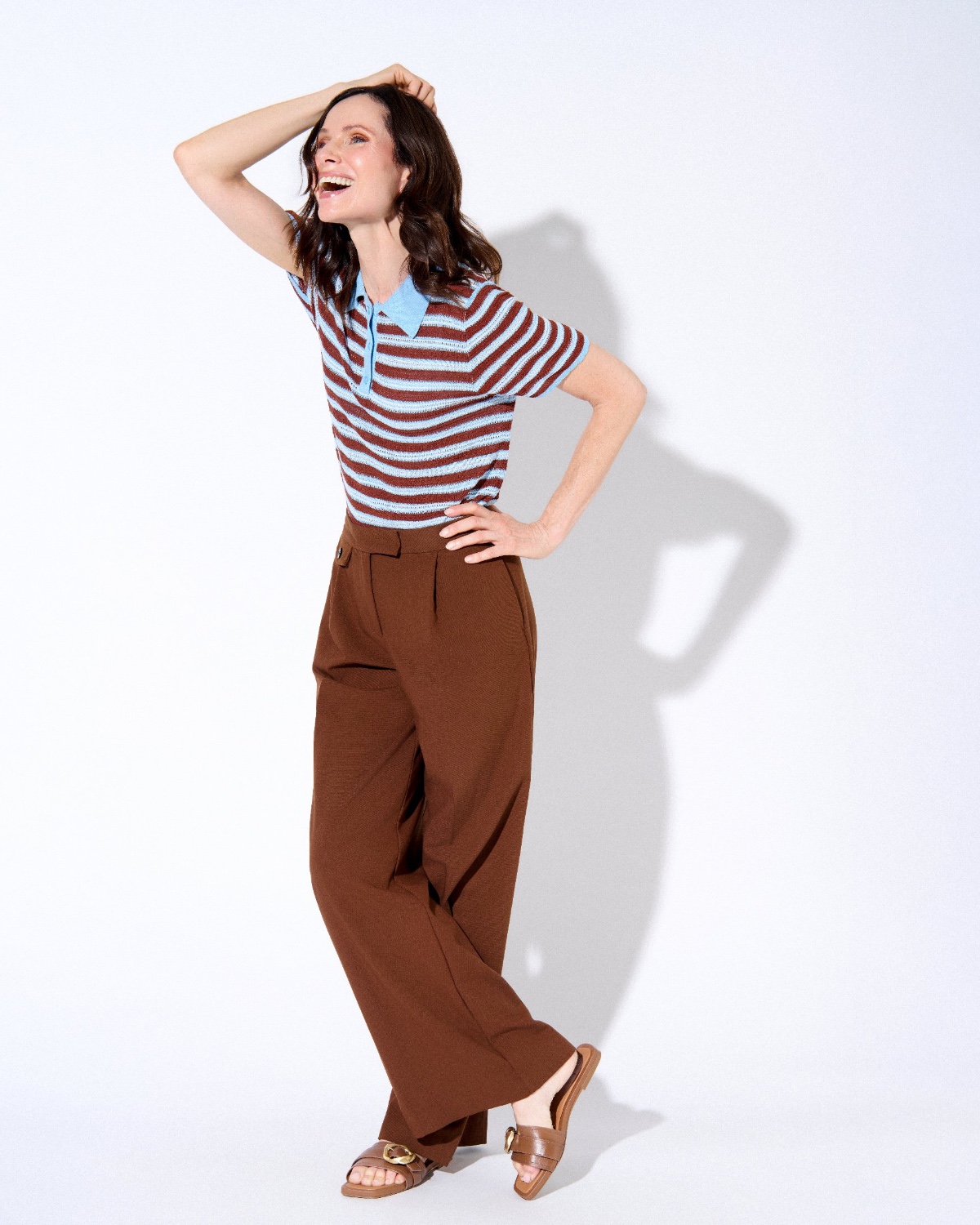 sasha-finn-12 A smiling woman in a striped polo and brown trousers poses against a bright white studio background.