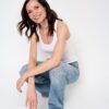 A smiling brunette woman in a white tank top and blue jeans poses against a white background.