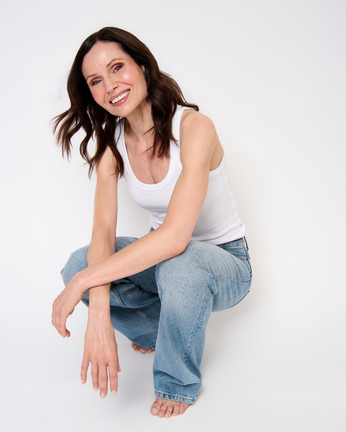 sasha-finn-18 A smiling brunette woman in a white tank top and blue jeans poses against a white background.