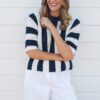 A smiling blonde woman wearing a beige sweater and white trousers sitting against a rustic wooden wall.