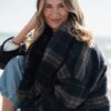 A smiling blonde woman wearing a warm plaid coat sits outdoors near the sea on a sunny day.