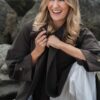 A smiling blonde woman wearing a brown coat sits outdoors on rocky terrain, looking happy and relaxed.