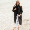A smiling blonde woman wearing a trench coat walks barefoot along the shoreline on a sandy beach.