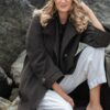 A smiling blonde woman sits on large rocks by the coast wearing a dark trench coat.